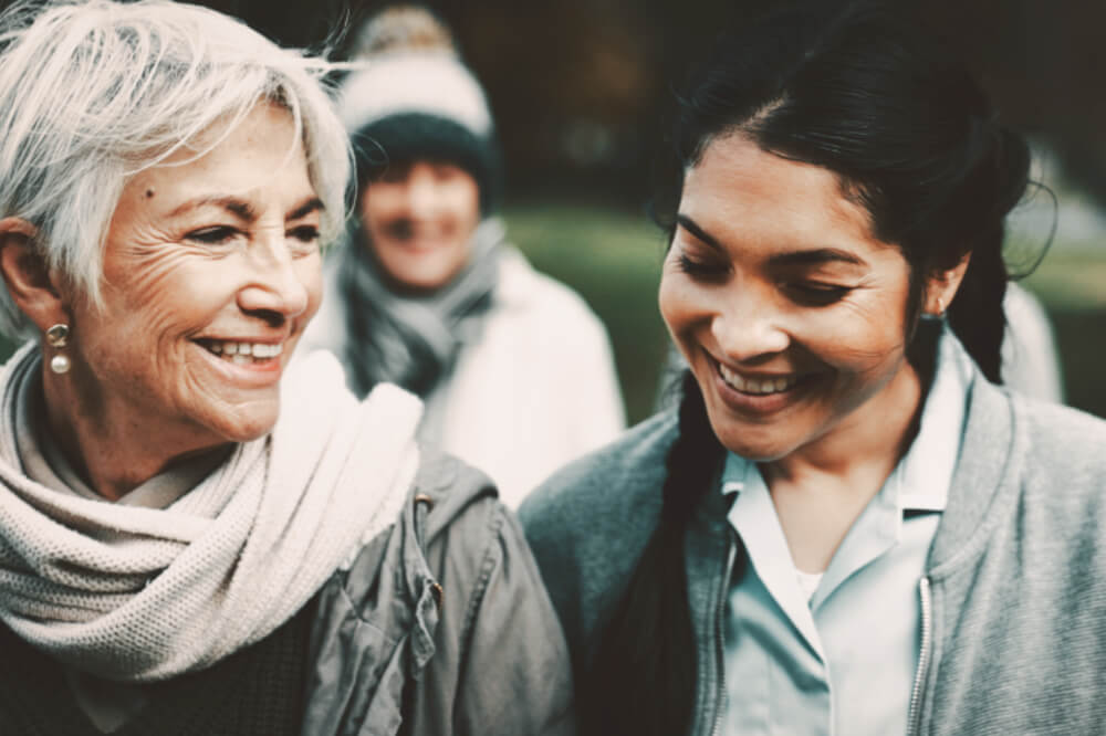 An elderly woman is walking alongside a younger woman, while the two chat and smile.