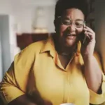 A person wearing glasses and a yellow shirt is talking on a phone while holding a cup, standing in a well-lit room.