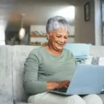 An elderly woman with short gray hair sits on a couch using a laptop, smiling while typing. The background shows a well-lit room with decor and bookshelves.