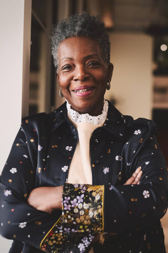 An elderly Black woman in a velvet jacket stands with her arms crossed.