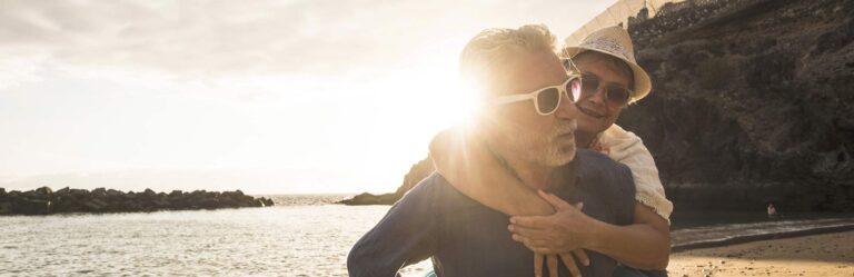 An elderly couple, both wearing sunglasses, are smiling on a beach at sunset. The man is giving the woman a piggyback ride. Rocks and water are visible in the background.