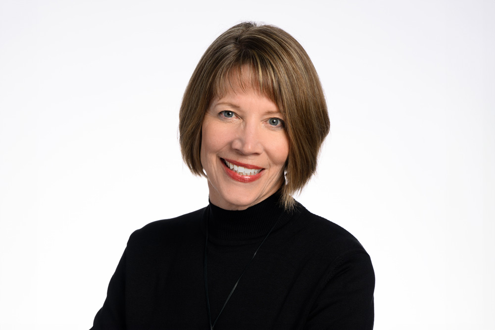 A woman with short, light brown hair smiles while wearing a black turtleneck against a plain, light background.