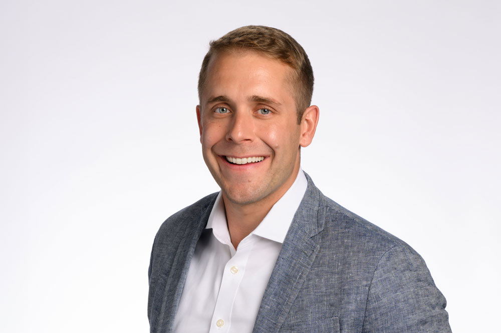 A man with short blonde hair and blue eyes is smiling and wearing a grey blazer over a white shirt against a plain white background.