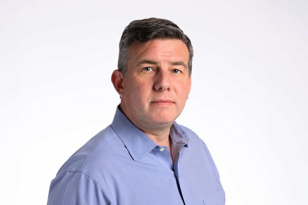 A man with short, dark hair wearing a light blue dress shirt stands against a plain white background.