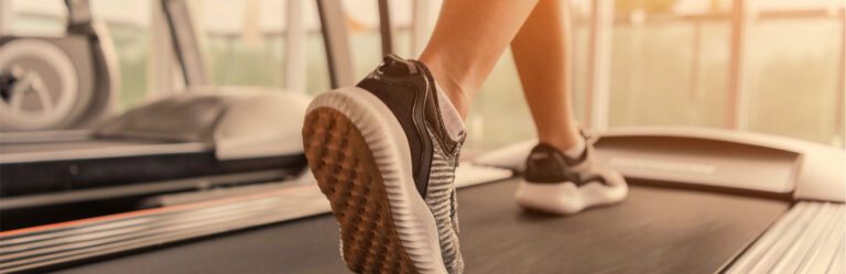 Close-up of a personâ€™s feet in sneakers walking on a treadmill, with gym equipment visible in the background.