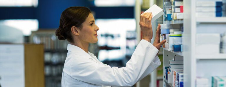 A person in a lab coat arranges boxes on a shelf in what appears to be a pharmacy or medical storage area.