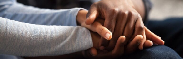 Close-up of two people holding hands, one with a light gray sleeve and the other with a dark skin tone.