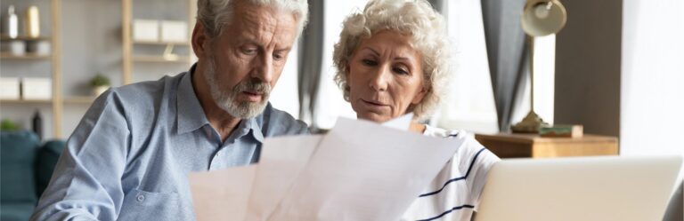 An elderly couple sits together, closely examining documents with a laptop in the foreground in a well-lit room.