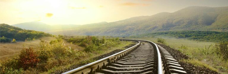 A railway track curves through a scenic landscape with rolling hills and greenery under a bright, hazy sky.