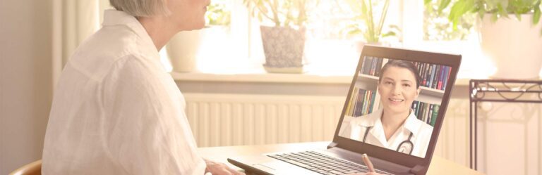 An elderly person in a white shirt participates in a video call with a doctor on a laptop in a bright room.