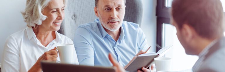 An elderly couple holding mugs is sitting across from a man discussing documents. The man is showing them something in a notebook.