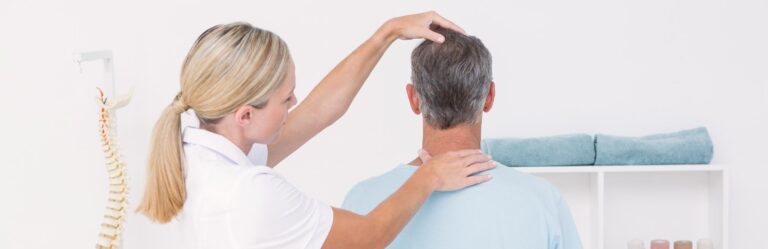 A female medical professional examines the neck and head of a seated male patient. A model of a spine and neatly folded towels are visible in the background.