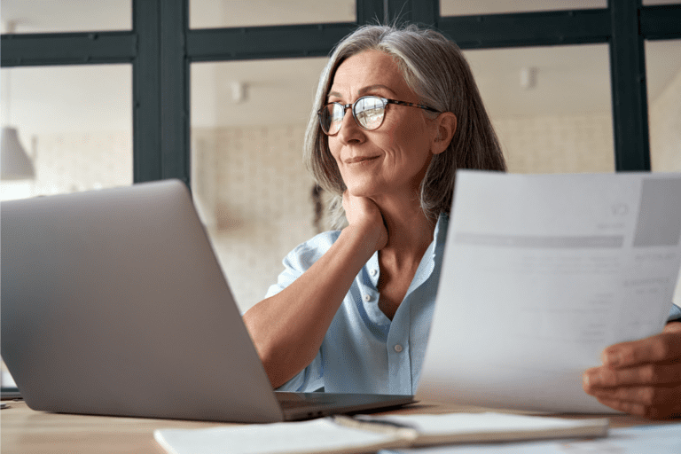 An older woman with gray hair and glasses sits at a desk, looking at her laptop while holding a document.