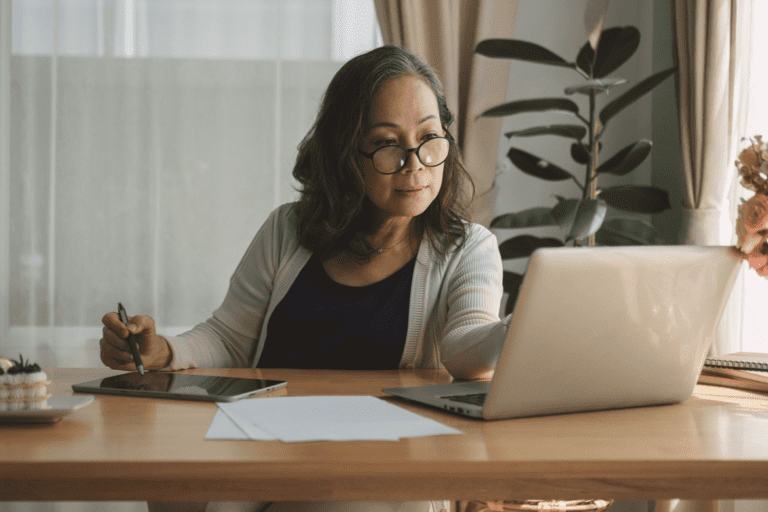 A person wearing glasses sits at a desk with a laptop, tablet, and documents, holding a stylus pen while working.