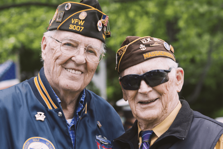 Two elderly military veterans wearing caps and jackets, standing side by side and smiling outdoors with trees in the background. One man is wearing glasses, and the other man is wearing sunglasses.