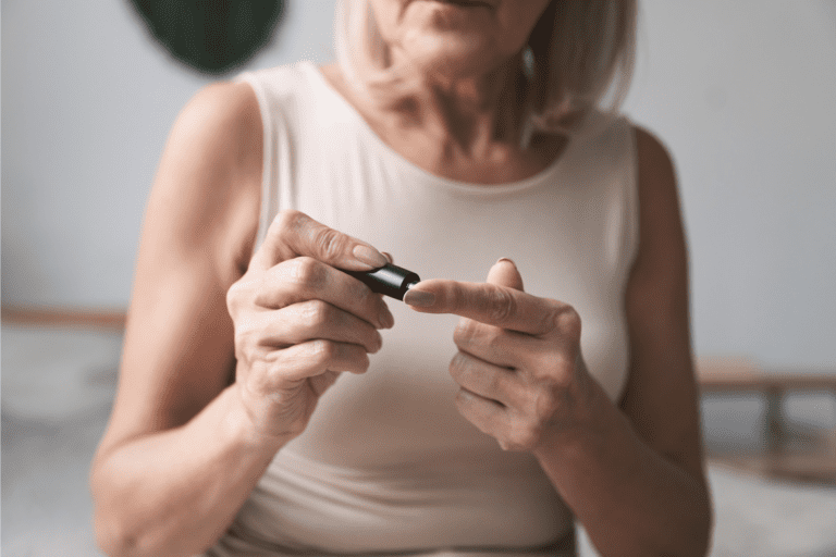 A person with light-colored hair uses a small device to check their blood glucose level by pricking their fingertip in a dimly lit room.