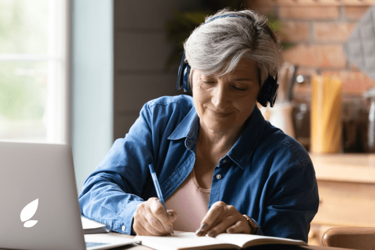 An older adult with short gray hair is writing in a notebook while wearing headphones and sitting at a desk with a laptop in a well-lit room.