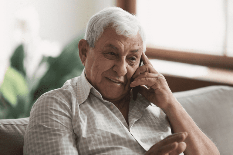 A senior man with white hair is sitting on a couch, talking on a mobile phone, and smiling.