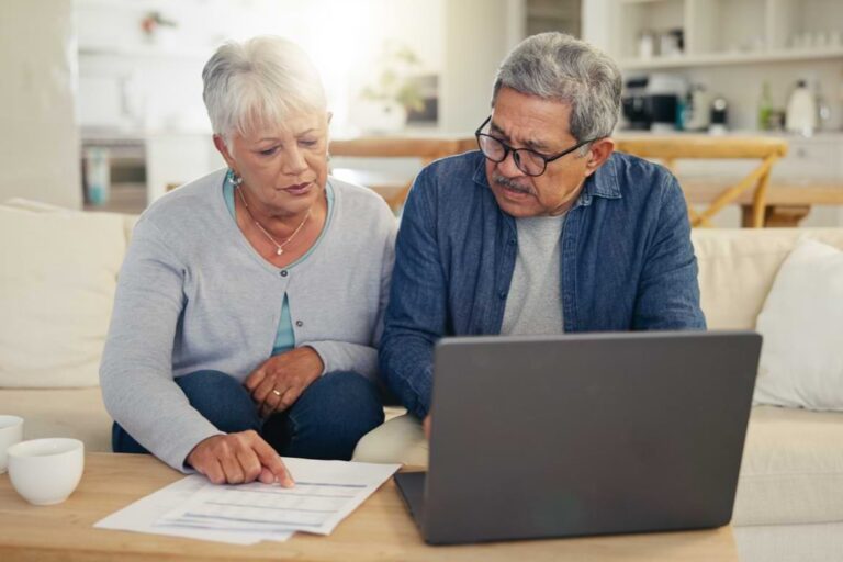 An older couple sits on a couch looking at a document and working on a laptop in a home setting.