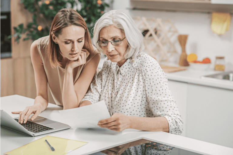 Two women, one younger and one older, review a document together in a kitchen. A laptop is open on the counter next to them.