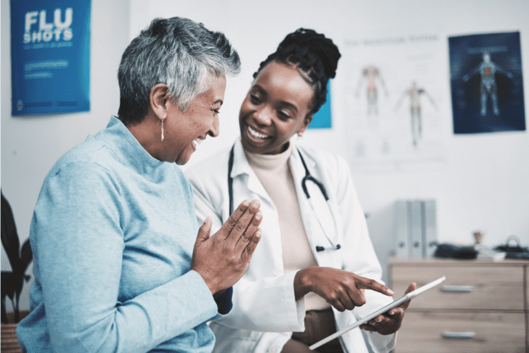 A doctor shows a tablet to an elderly patient in a medical office. Both are smiling.