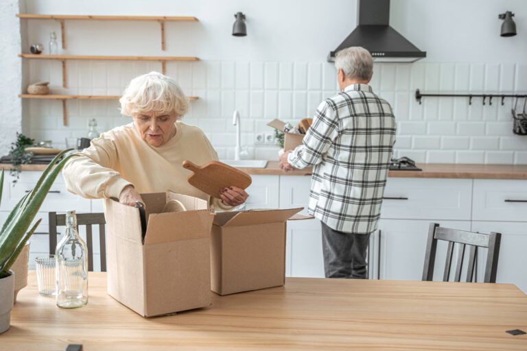 An elderly woman and man unpack boxes in a modern kitchen with white cabinets and a wooden table.