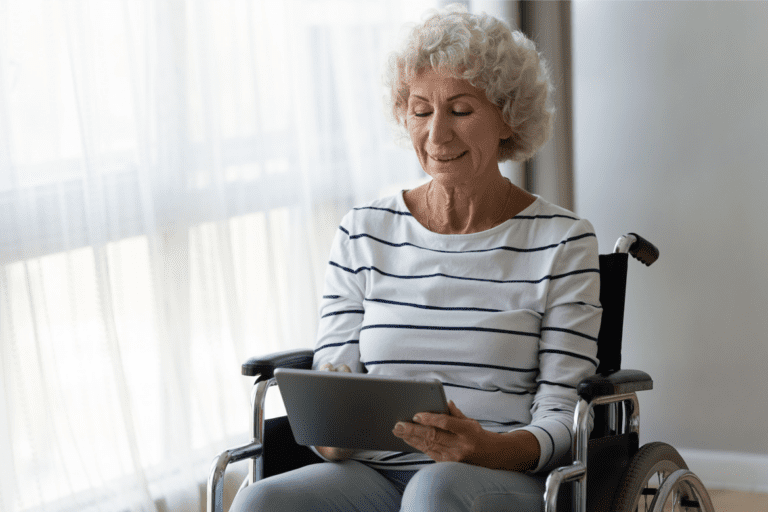 An elderly woman with curly gray hair sits in a wheelchair near a window, looking at a tablet. She is wearing a white and blue striped shirt and appears to be focused on the device.