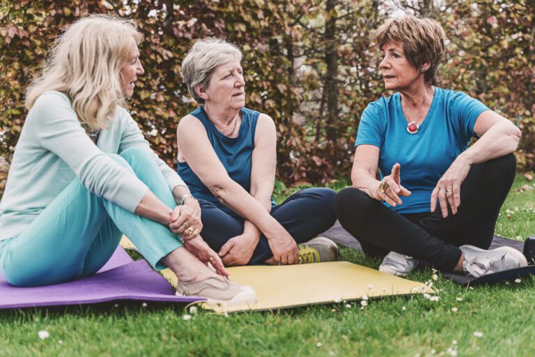 Three women sit on yoga mats in a grassy outdoor area, engaged in conversation.