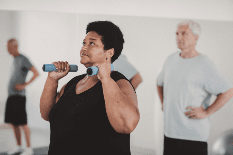 A person is standing in a gym holding blue dumbbells at shoulder height, while another person stands in the background with hands on hips.