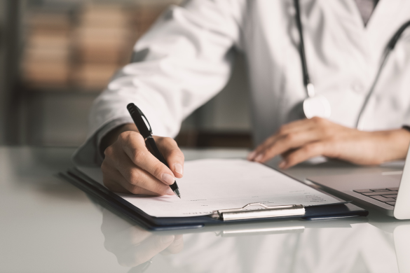 A medical professional in a white coat writes on a clipboard with a pen, with a stethoscope around their neck and a laptop on the desk.