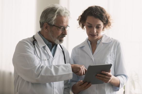 Two healthcare professionals are discussing something displayed on a tablet. One is an older man with grey hair and glasses, wearing a stethoscope. The other is a younger woman with curly hair.