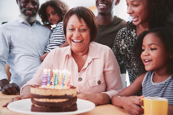 A woman is seated at a table with a cake topped with lit candles while people around her smile and celebrate.