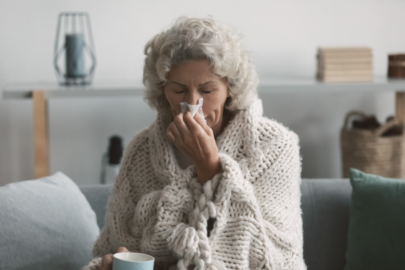 An elderly woman with gray hair sits on a couch, wrapped in a thick knitted blanket, holding a teacup, and blowing her nose into a tissue.