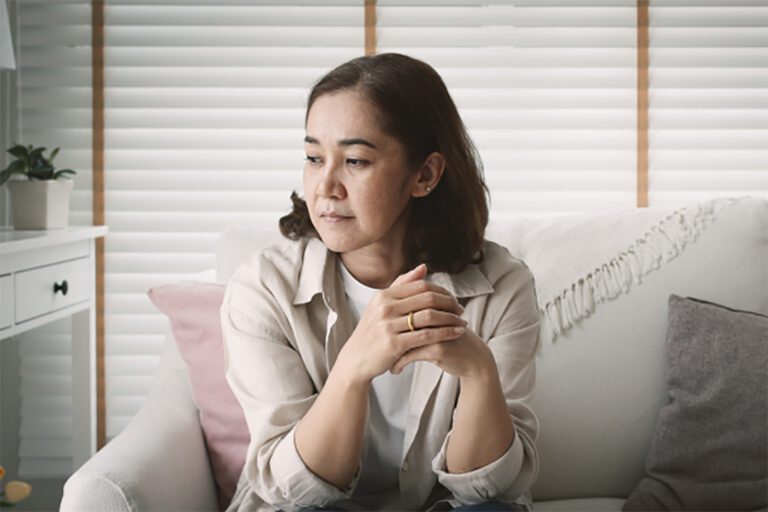 A woman sits on a white couch in a living room with white blinds, looking thoughtfully to the side with her hands clasped.