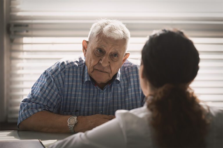 An elderly man with white hair sits across from a person with dark hair, visible from the back, having a conversation in a room with horizontal blinds.