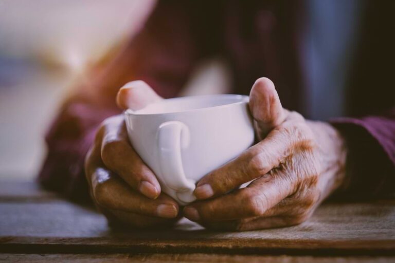 Close-up of an elderly person's hands holding a white mug, resting on a wooden table. The individual is wearing a maroon long-sleeve shirt. The background is softly blurred.