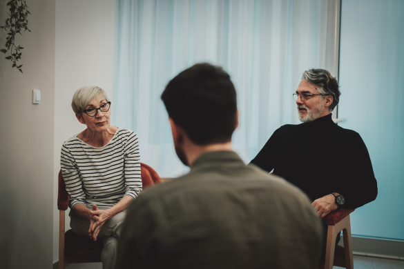 Two older adults, a woman and a man, sit on chairs facing a younger man, who is sitting with his back to the camera in a room with sheer curtains.