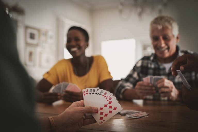 Three people are playing cards at a wooden table in a bright room. The focus is on a hand holding playing cards, showing a combination of cards.