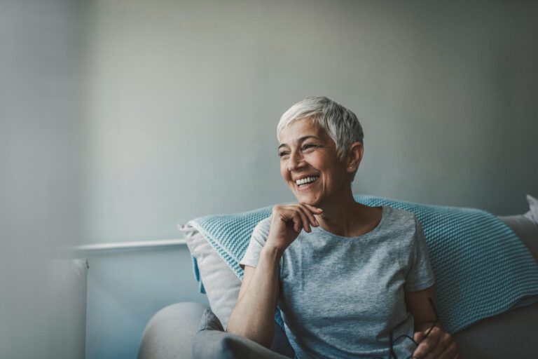 A woman with short gray hair and a gray t-shirt sits on a couch, smiling and looking to the side. She holds a pair of glasses in her left hand. A blue blanket is draped over the couch.