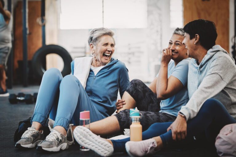 Three elderly women in workout clothes sit on a gym floor, laughing and engaging in conversation with water bottles nearby.