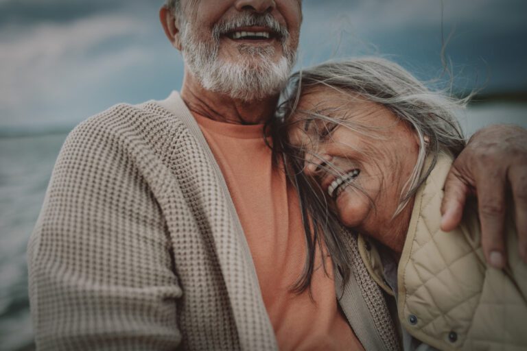 An elderly couple smiles while embracing, with the woman resting her head on the man's chest. They are outdoors with a cloudy sky in the background.