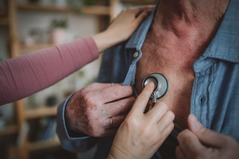 A person uses a stethoscope to listen to the chest of another person wearing an open shirt, with a hand resting on their shoulder.