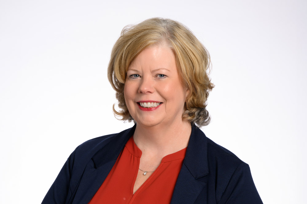 A woman with short blonde hair and a smile is wearing a red blouse and dark blazer, standing against a white background.