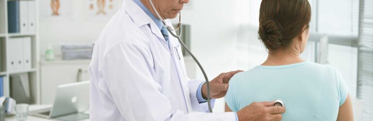 A doctor using a stethoscope on a patient's back during a medical examination in an office setting.