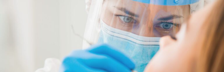 A healthcare worker wearing a mask, face shield, and gloves examines a patient's mouth.