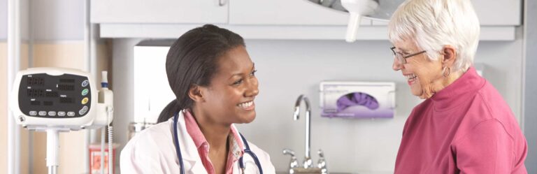 A doctor smiles and speaks with an elderly patient in a medical office. A blood pressure monitor is visible in the background.