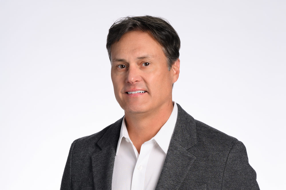 A man with dark hair wearing a white dress shirt and a gray blazer stands against a plain white background.