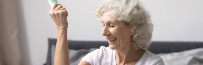 A smiling elderly person with short, curly gray hair holds up a tube of lotion while sitting indoors, dressed in a white top.