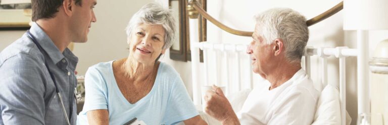 A young man in a denim shirt converses with an elderly woman and an elderly man sitting up in bed in a warmly lit room. The elderly man holds a cup.