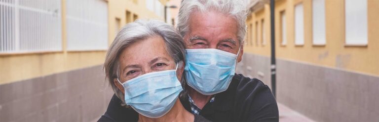 Two elderly individuals wearing face masks stand close together in an outdoor setting between buildings, embracing each other.